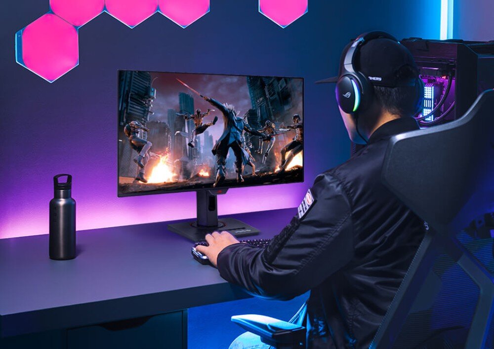 Osoba siedzi przy biurku, grając na komputerze z dużym monitorem, na którym wyświetlana jest dynamiczna scena z gry. W tle widoczne są różowe, sześciokątne panele świetlne na ścianie.