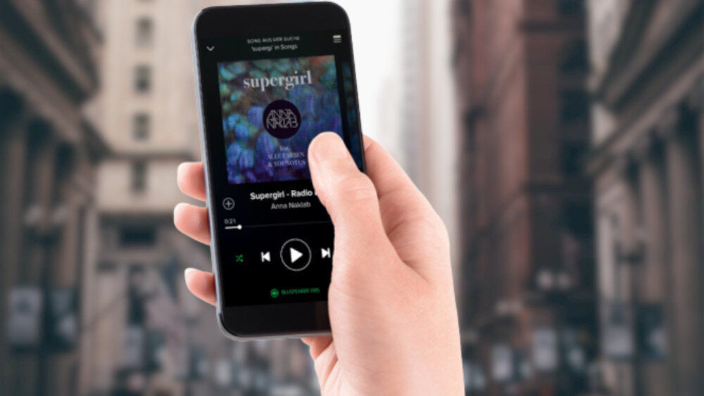 Osoba trzyma smartfon, na którym wyświetlana jest aplikacja muzyczna z utworem \'Supergirl - Radio Edit\' autorstwa Anna Naklab. W tle widać rozmyty obraz miejskiej ulicy z wysokimi budynkami.