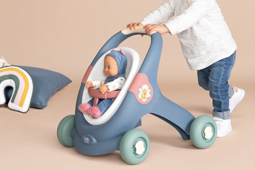 Dziecko pcha niebieski wózek z lalką w środku, na wózku widnieje napis \'Little Smoby\'. Obok znajduje się poduszka w kształcie tęczy.