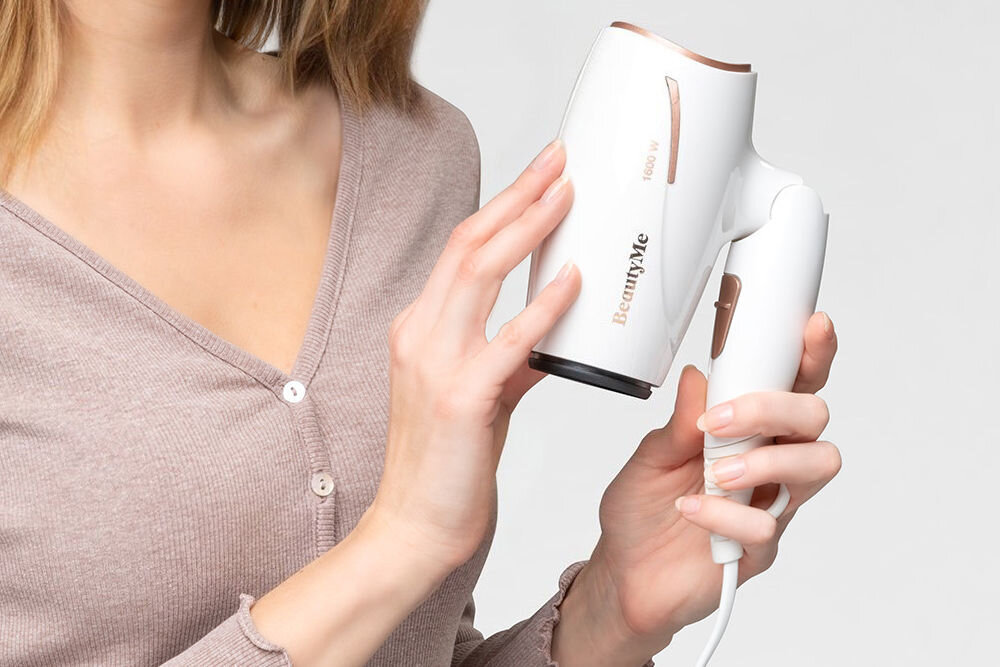 Osoba trzyma białą suszarkę do włosów z napisem \'BeautyMe\' i oznaczeniem \'1600 W\'. Suszarka ma złote detale i jest podłączona do prądu.