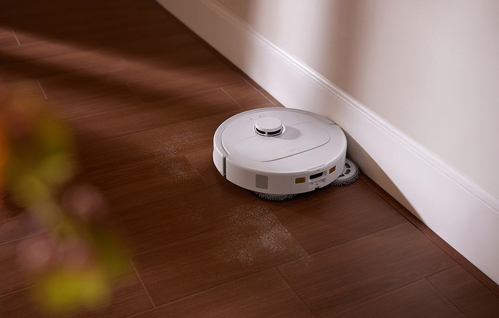 Biały robot sprzątający porusza się po drewnianej podłodze wzdłuż ściany. Na podłodze widoczny jest ślad kurzu, który robot sprząta.