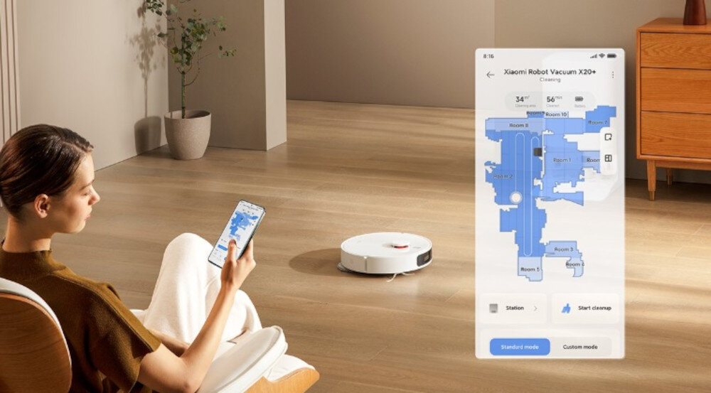 Osoba siedzi na krześle, trzymając w ręku smartfon z aplikacją pokazującą mapę sprzątania. Na podłodze widać biały robot sprzątający.