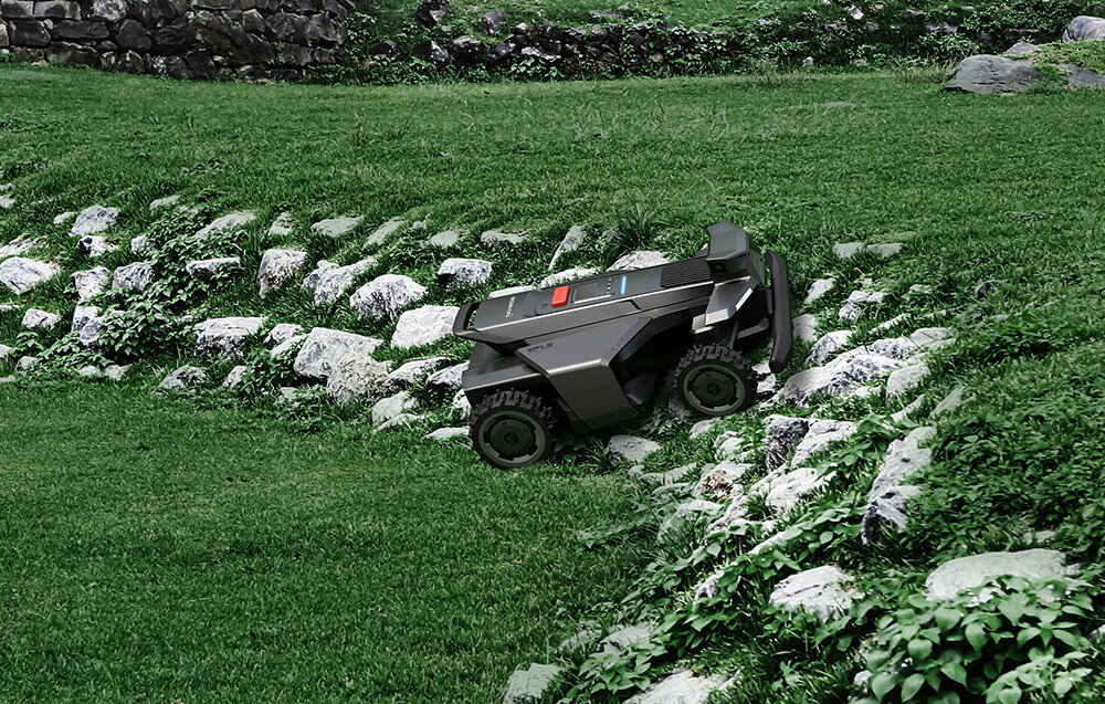 Robot koszący porusza się po trawiastym terenie z kamienistymi elementami. Urządzenie ma ciemny kolor i widoczne są na nim dwa kolorowe panele.