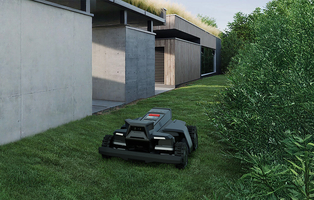 Na trawniku przed nowoczesnym budynkiem znajduje się robot koszący. W tle widoczne są betonowe i drewniane elementy architektury oraz roślinność.