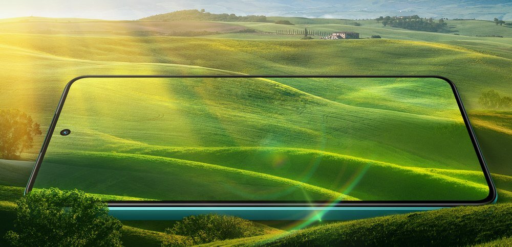 Smartfon z dużym ekranem wyświetla krajobraz z zielonymi wzgórzami i promieniami słońca. Obraz na ekranie płynnie łączy się z tłem, tworząc iluzję ciągłości.