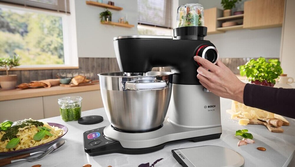 Na blacie kuchennym znajduje się robot kuchenny z metalową misą i pokrętłem, obok którego widoczna jest dłoń. W tle widać kuchenne akcesoria i składniki, takie jak makaron i zioła.
