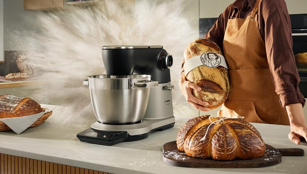 Na blacie kuchennym stoi robot kuchenny z metalową misą, obok osoba trzymająca duży bochenek chleba. W tle widać rozpryskującą się mąkę, a na blacie leży chleb na desce do krojenia.