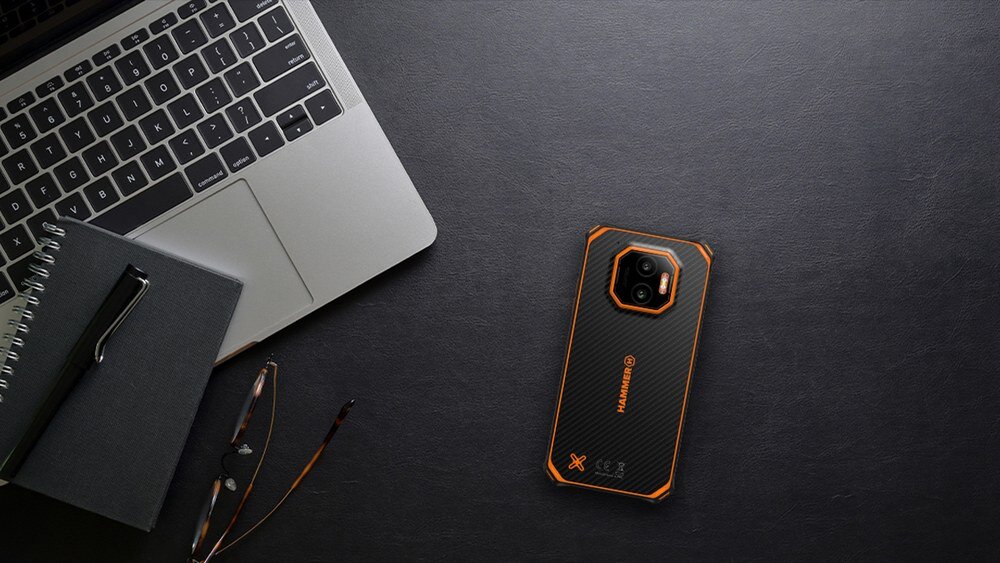 Na ciemnym biurku leży smartfon z czarno-pomarańczową obudową obok otwartego laptopa, notatnika z długopisem i okularów. Na obudowie telefonu widoczne jest logo \'HAMMER\'.