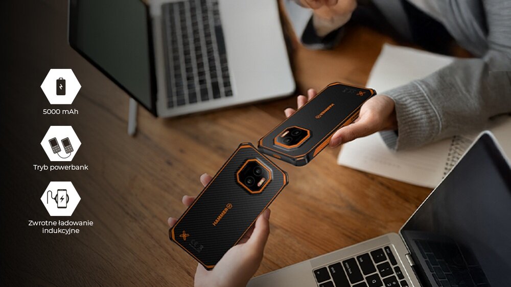 Dwie osoby trzymają smartfony o czarno-pomarańczowym designie nad drewnianym stołem, obok laptopów. Po lewej stronie widoczne są ikony z tekstem: \'5000 mAh\', \'Tryb powerbank\', \'Zwrotne ładowanie indukcyjne\'.