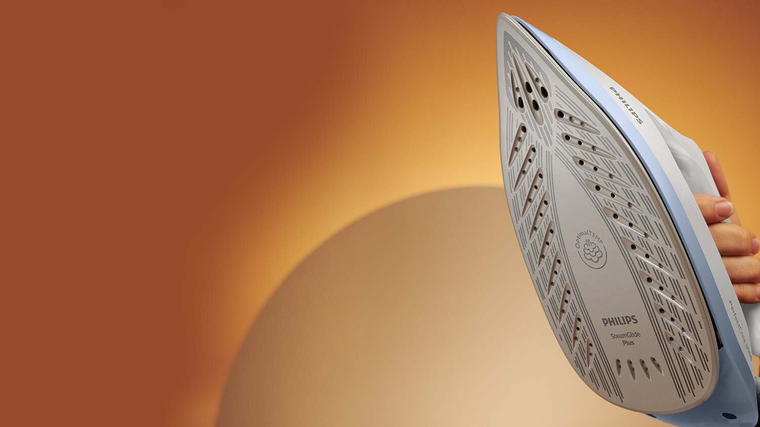 Zdjęcie przedstawia stopę żelazka z widocznymi otworami parowymi i napisem \'PHILIPS SteamGlide Plus\'. W tle znajduje się rozmyte, pomarańczowe tło.