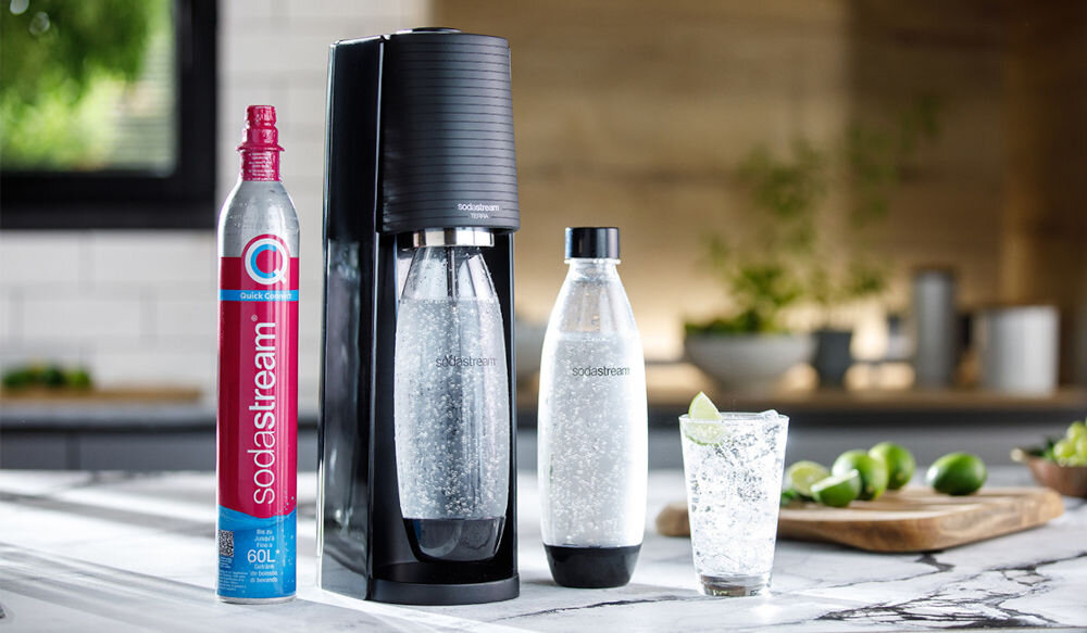 Na blacie kuchennym stoi czarny saturator z przezroczystą butelką wypełnioną gazowaną wodą. Obok znajduje się butla z napisem \'Sodastream\' oraz szklanka z napojem i plasterkiem limonki.