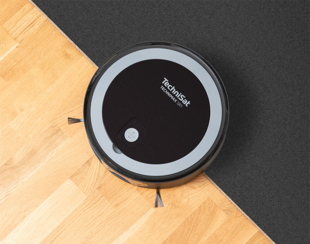 Na obrazie widoczny jest okrągły robot sprzątający na granicy dwóch powierzchni: drewnianej podłogi i ciemnego dywanu. Na robocie znajduje się napis \'TechniSat TECHNIMAX SR1\'.