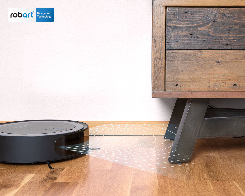 Robot sprzątający porusza się po drewnianej podłodze obok drewnianej szafki. W lewym górnym rogu widoczne jest logo \'robart Navigation Technology\'.