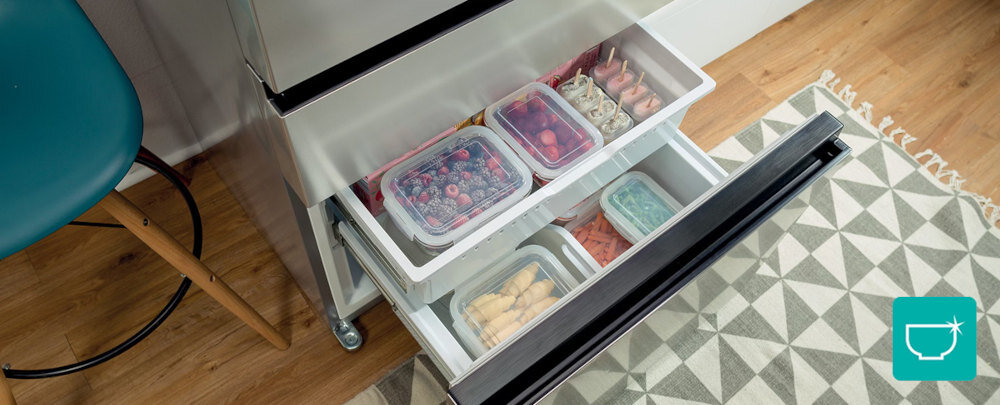 Otwarta szuflada lodówki z widocznymi pojemnikami na żywność, wypełnionymi owocami i innymi produktami. Obok lodówki znajduje się krzesło i dywan z geometrycznym wzorem.