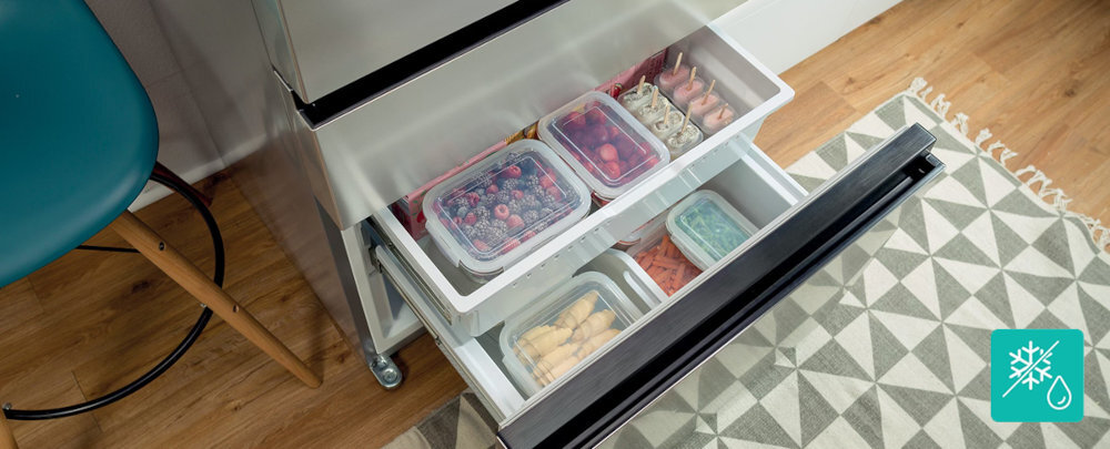Otwarta szuflada lodówki z widocznymi pojemnikami na żywność, wypełnionymi owocami i innymi produktami. Obok lodówki znajduje się niebieskie krzesło i dywan z geometrycznym wzorem.