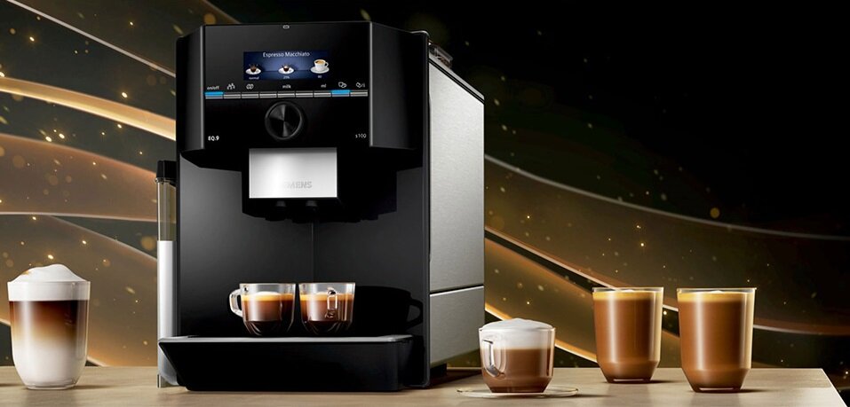 Czarny ekspres do kawy z wyświetlaczem, na którym widoczny jest napis \'Espresso Macchiato\'. Obok ekspresu znajdują się filiżanki z różnymi rodzajami kawy.
