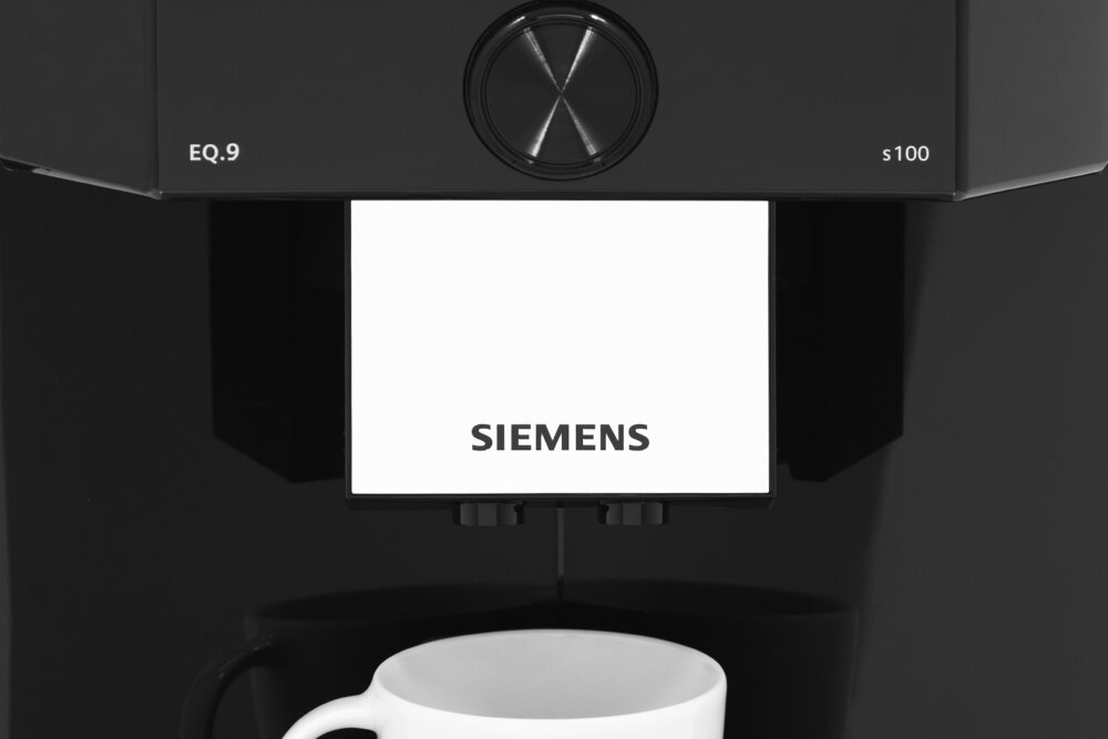 Czarny ekspres do kawy z widocznym logo \'SIEMENS\' na białym tle. Pod dyszami znajduje się biała filiżanka.