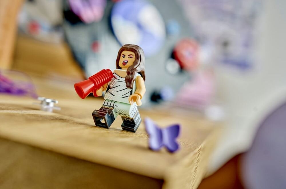 Figurka LEGO przedstawiająca postać z długimi włosami trzymająca czerwony megafon. W tle widoczny jest rozmyty obraz z elementami w różnych kolorach.