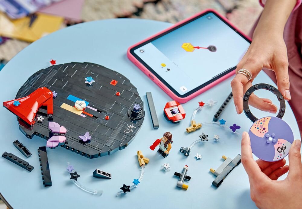 Na okrągłym, czarnym obiekcie z klocków LEGO znajdują się kolorowe elementy i ozdoby, obok leży tablet z aplikacją. Widać także ręce osoby trzymającej część konstrukcji LEGO oraz rozrzucone klocki na stole.