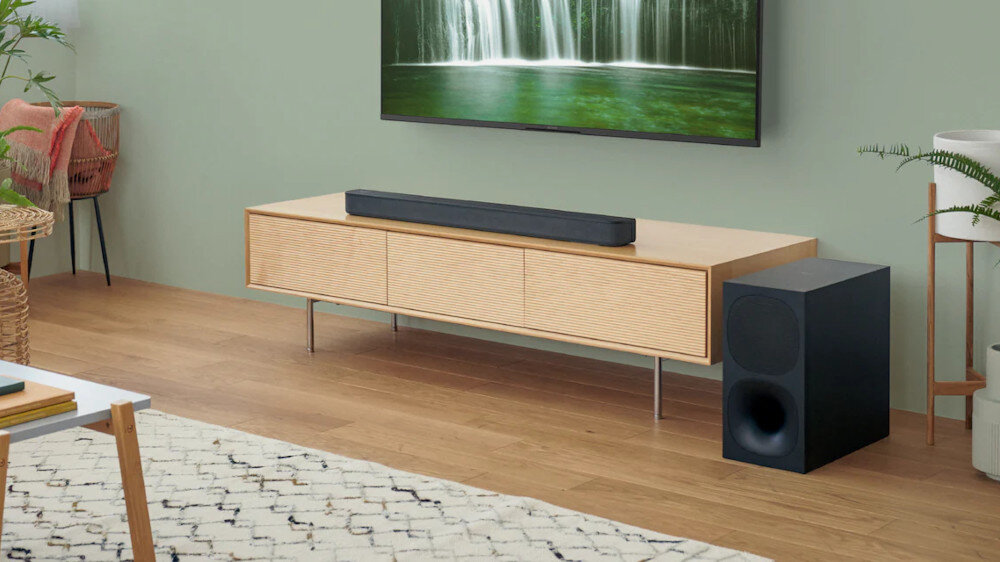 Na drewnianej szafce pod telewizorem znajduje się czarny soundbar, a obok stoi czarny subwoofer. W tle widać telewizor z obrazem wodospadu.