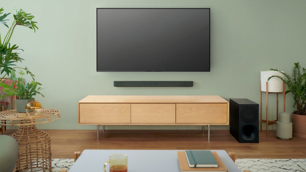 Na obrazie znajduje się nowoczesny salon z telewizorem zamontowanym na ścianie, poniżej którego umieszczony jest czarny soundbar. Obok drewnianej szafki znajduje się czarny subwoofer, a w pomieszczeniu widoczne są rośliny i stolik z książką.