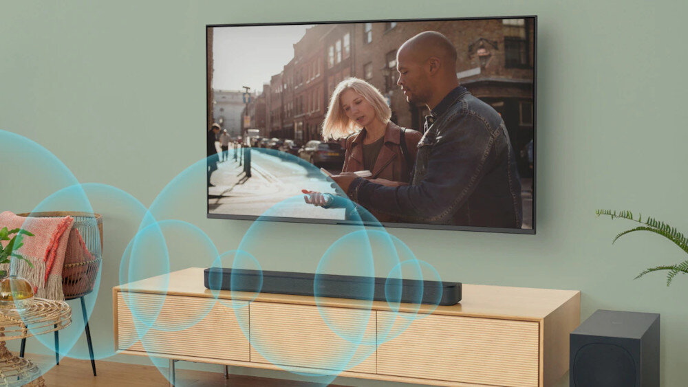 Na drewnianej szafce stoi czarny soundbar, przed którym widoczne są niebieskie fale dźwiękowe. Na ścianie wisi telewizor z obrazem przedstawiającym dwie osoby na ulicy.