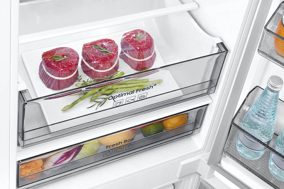 Wnętrze lodówki z szufladą oznaczoną jako \'Optimal Fresh+\', w której znajdują się trzy kawałki mięsa i kilka szparagów. Poniżej widoczna jest szuflada \'Fresh Box\' z owocami i warzywami, a na drzwiach butelki z wodą.