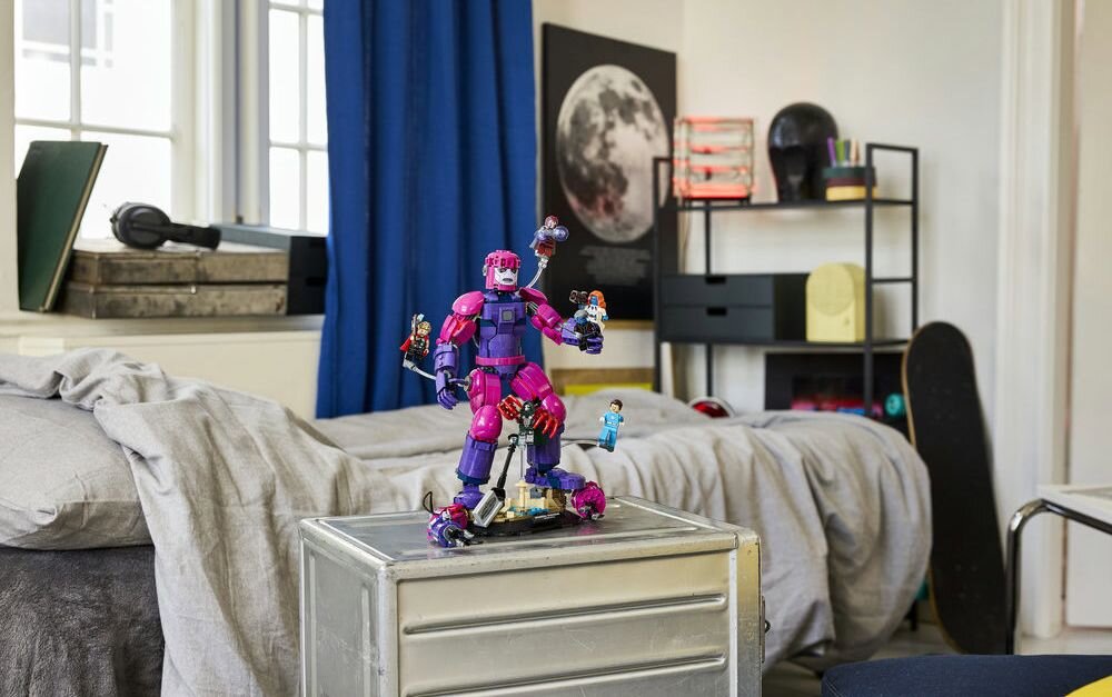 Na metalowej szafce stoi duża figurka robota w kolorach fioletowym i różowym, otoczona mniejszymi figurkami. W tle widać łóżko z szarą pościelą, niebieską zasłonę i plakat z księżycem na ścianie.