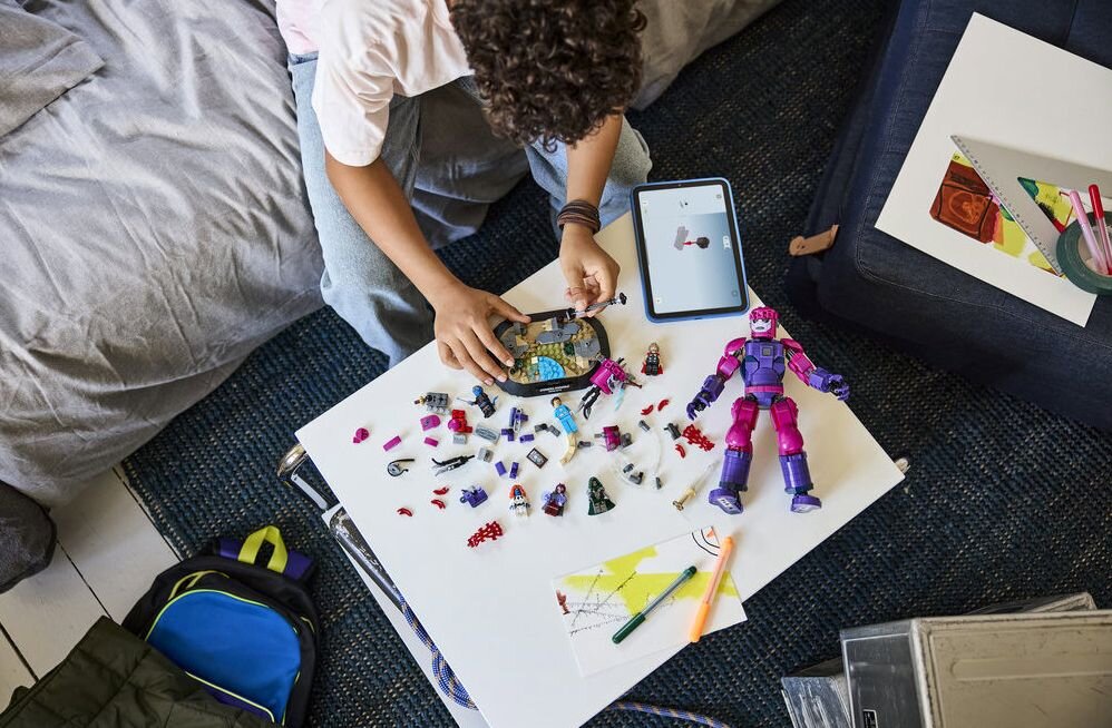 Na białym stole znajdują się rozłożone elementy zestawu LEGO oraz tablet z wyświetlonym obrazem. Obok leży duża, fioletowo-różowa figurka robota.