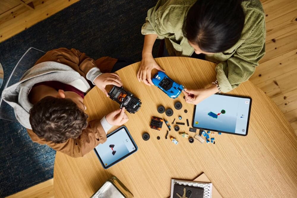 Na okrągłym stole znajdują się dwa modele samochodów LEGO, kilka klocków oraz dwa tablety z wyświetlonymi instrukcjami. Dwie osoby składają modele, a wokół leżą różne elementy zestawu LEGO.