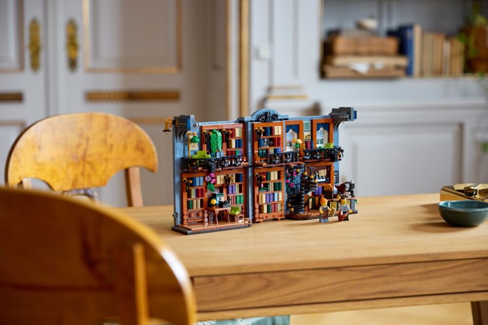 Na drewnianym stole znajduje się zestaw LEGO przedstawiający miniaturową księgarnię z detalami takimi jak półki z książkami i małe figurki. W tle widoczne są eleganckie drzwi i półki z książkami w jasnym pomieszczeniu.