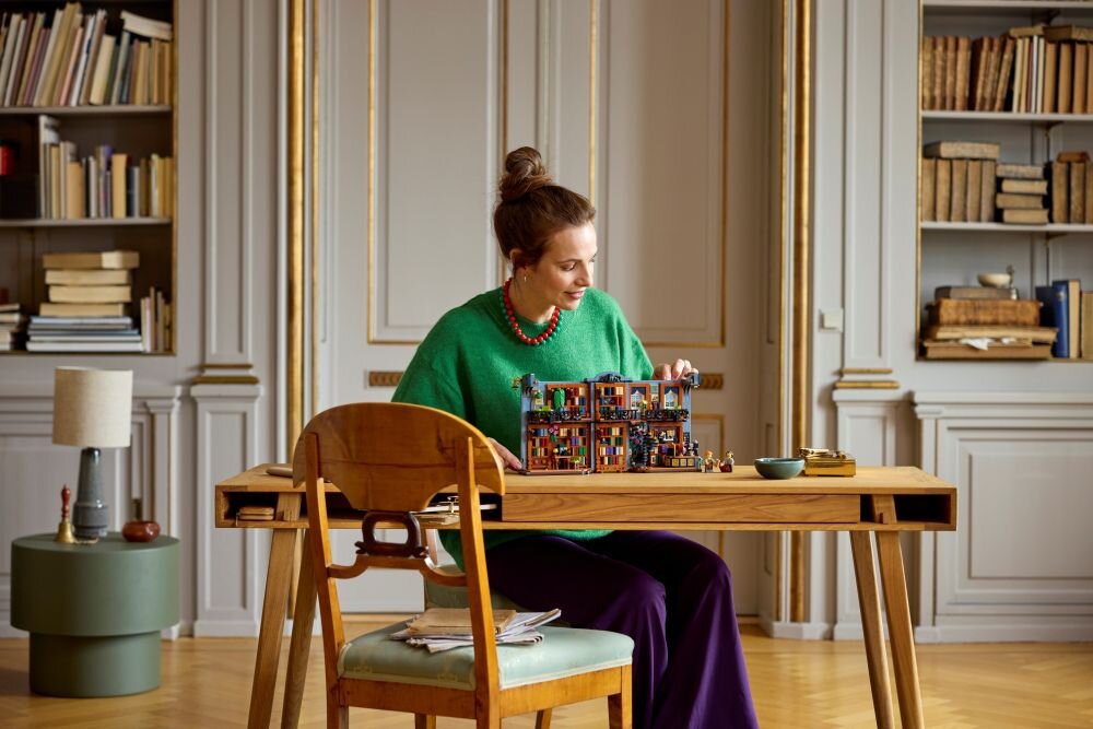 Osoba siedzi przy drewnianym stole, na którym znajduje się złożony zestaw LEGO przedstawiający księgarnię. W tle widać regały z książkami i eleganckie, klasyczne wnętrze.