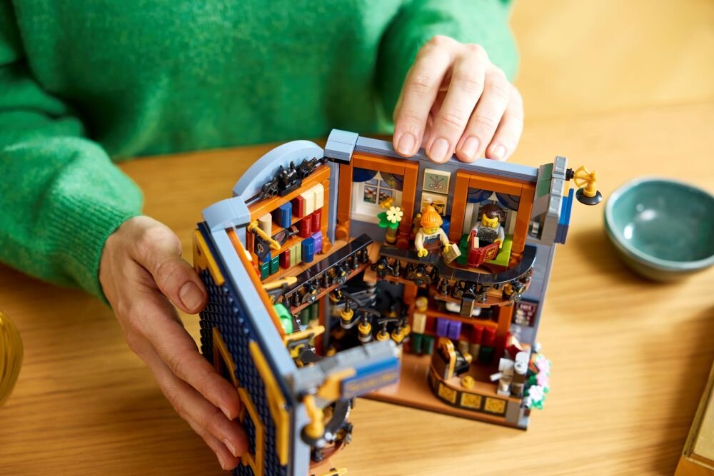 Osoba w zielonym swetrze trzyma model LEGO przedstawiający wnętrze księgarni z półkami pełnymi książek i figurkami. Na stole obok znajduje się mała miska.