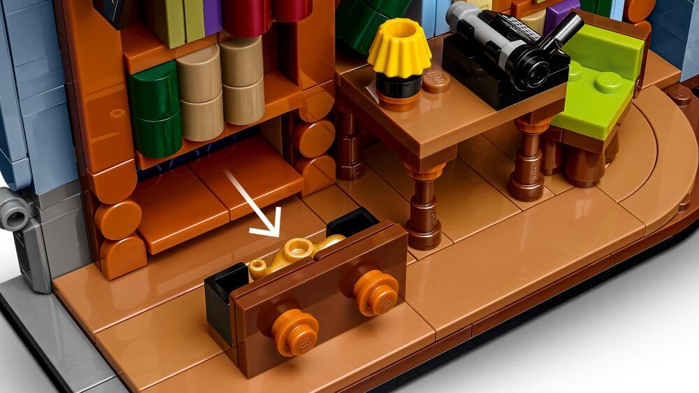 Zestaw LEGO przedstawiający wnętrze księgarni z półkami pełnymi książek, biurkiem z lampką i maszyną do pisania oraz szufladą z klockami. Na podłodze widoczna jest biała strzałka wskazująca na otwartą szufladę.