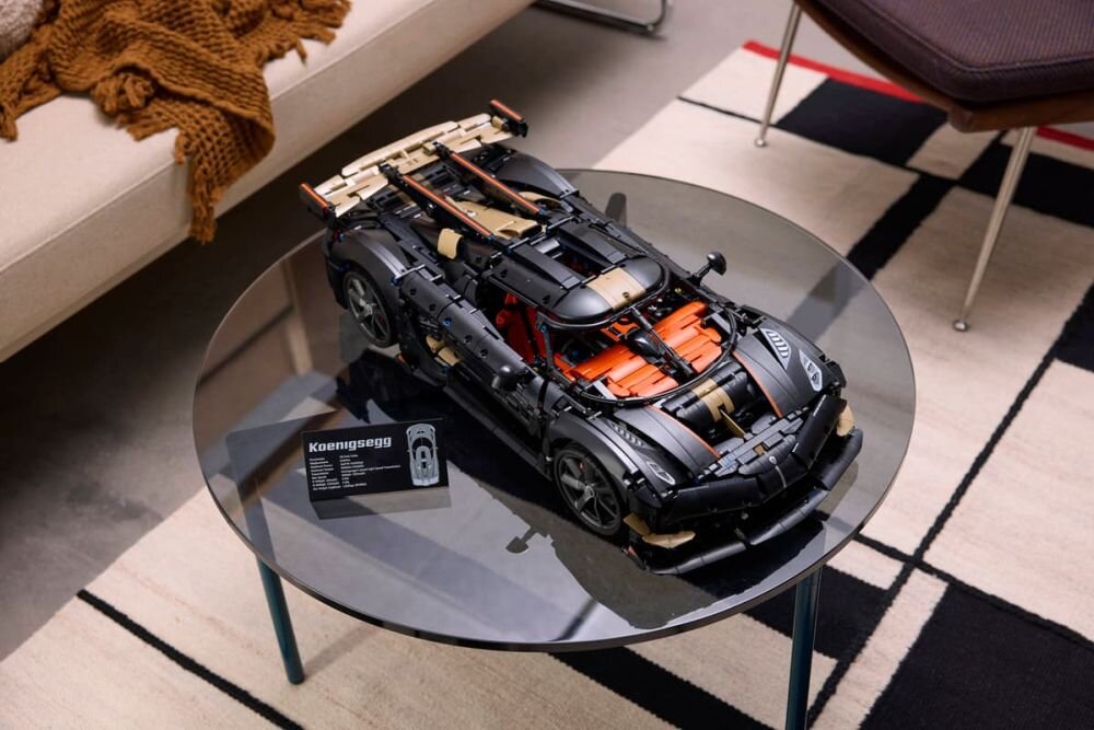 Na szklanym stoliku znajduje się model samochodu z klocków LEGO, obok którego stoi tabliczka z napisem \'Koenigsegg\'. W tle widoczna jest część kanapy i dywanu z geometrycznym wzorem.