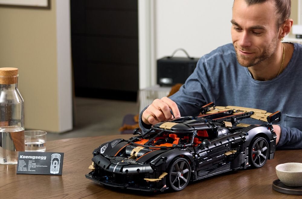 Osoba składa model samochodu z klocków LEGO na stole, obok stoi karafka z wodą i szklanka. Na stole znajduje się również tabliczka z napisem \'Koenigsegg\'.