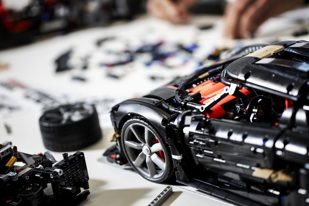 Na obrazie widoczny jest model samochodu z klocków LEGO, z wyraźnie zaznaczonymi elementami technicznymi i kołem. W tle znajdują się rozłożone części LEGO oraz dłonie osoby składającej model.