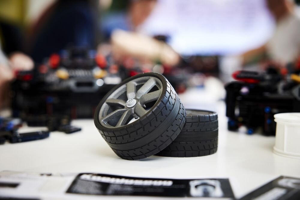 Na stole znajdują się dwa czarne koła z szarymi felgami, prawdopodobnie elementy zestawu LEGO. W tle widoczne są rozmyte sylwetki osób oraz inne części konstrukcyjne.