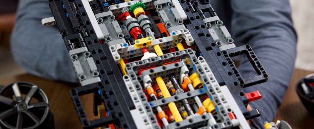 Zbliżenie na konstrukcję z klocków LEGO Technic, przedstawiającą skomplikowany mechanizm z widocznymi elementami w kolorach czarnym, żółtym, czerwonym i szarym. Obok znajduje się koło z czarną felgą.