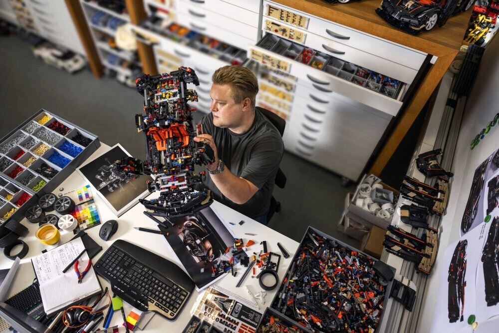 Osoba siedzi przy biurku, trzymając w rękach złożony model LEGO, otoczona różnymi elementami LEGO i narzędziami. Na biurku znajdują się także klawiatura, myszka i notatki.