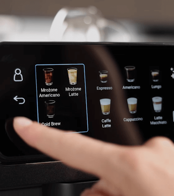 Na ekranie dotykowym ekspresu do kawy widoczne są opcje napojów, takie jak \'Mrożone Americano\', \'Mrożone Latte\', \'Cold Brew\', \'Espresso\', \'Americano\', \'Lungo\', \'Caffe Latte\', \'Cappuccino\' i \'Latte Macchiato\'. Palec wskazuje na opcję \'Cold Brew\'.