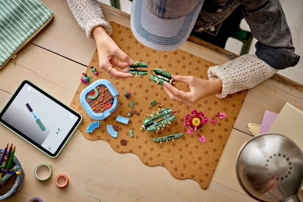 Osoba składa zestaw klocków LEGO na drewnianym stole, obok leży tablet z instrukcją. Na macie znajdują się różnokolorowe elementy, w tym zielone części przypominające kaktusy.