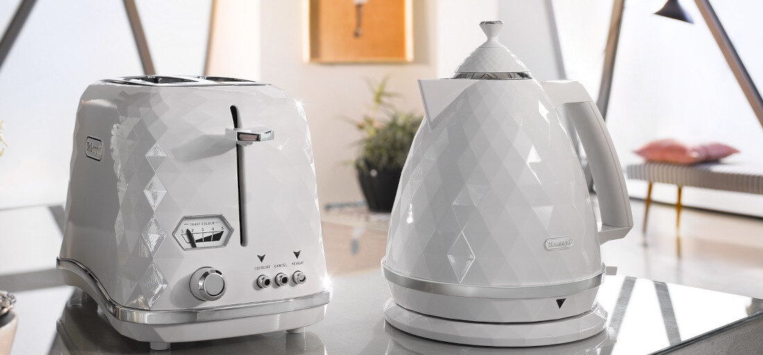 Na obrazie widoczny jest biały czajnik o geometrycznym wzorze oraz biały toster z podobnym wzorem. Oba urządzenia mają logo \'DeLonghi\'.