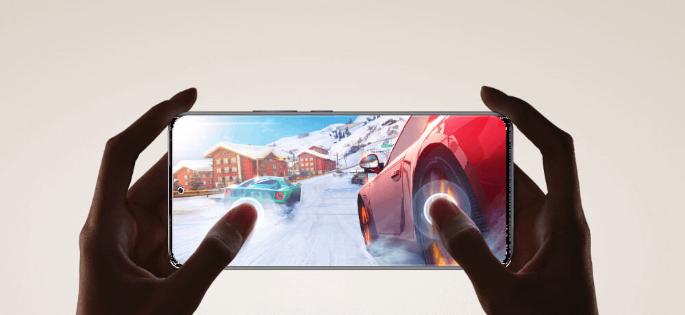 Osoba trzyma smartfon, na którym wyświetlana jest gra wyścigowa z samochodami na zaśnieżonej drodze. W tle widoczne są góry i budynki pokryte śniegiem.