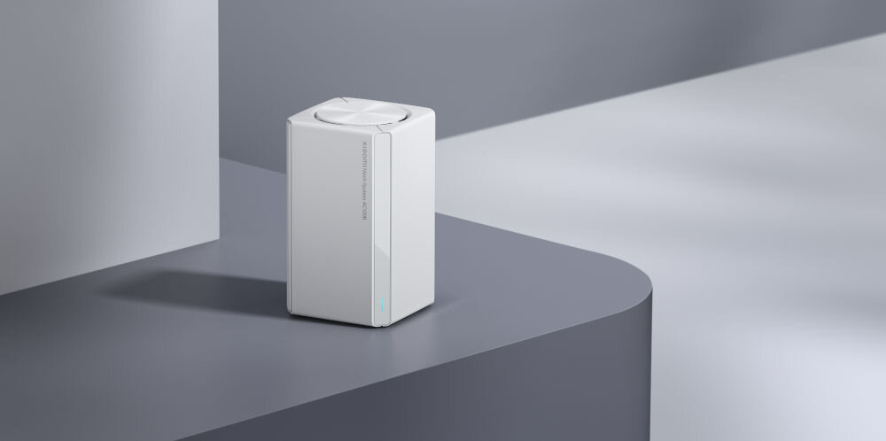 Na obrazie widoczny jest biały, prostokątny router z napisem na boku. Router stoi na szarej, gładkiej powierzchni.