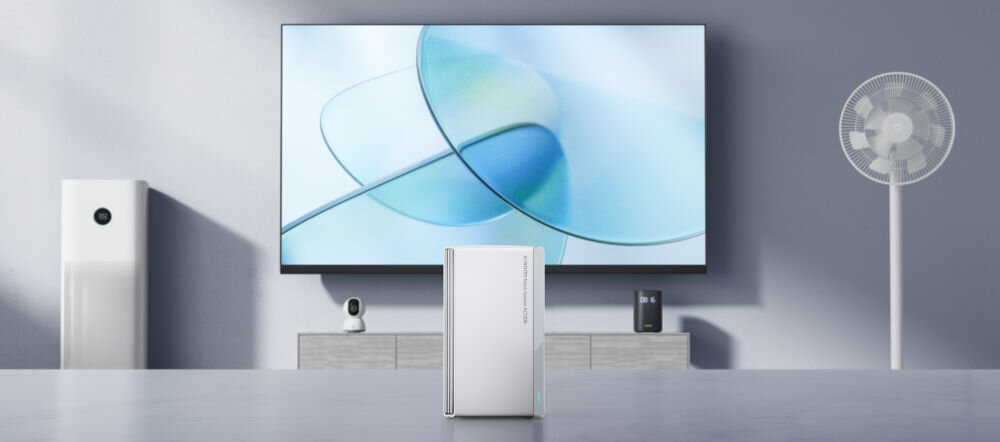 Na pierwszym planie znajduje się biały router o prostokątnym kształcie. W tle widoczny jest duży telewizor z abstrakcyjną grafiką, wentylator stojący oraz inne urządzenia elektroniczne na szafce.