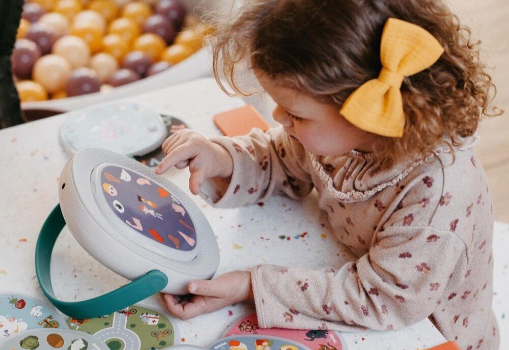 Dziecko bawi się interaktywną zabawką TIMIO Play Learn & Explore, która ma okrągły kształt i kolorowy panel z ilustracjami. Na stole wokół zabawki leżą różne kolorowe dyski.