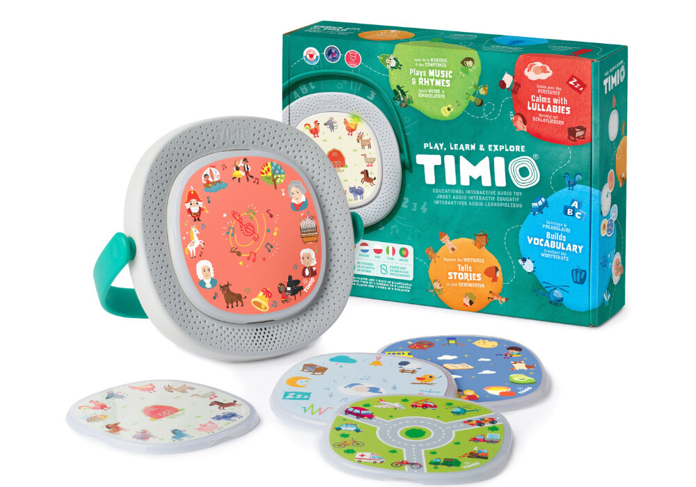 Na obrazie znajduje się interaktywna zabawka TIMIO z kolorowymi dyskami oraz opakowanie z napisem \'Play, Learn & Explore TIMIO\'. W tle widoczne są różne ilustracje i napisy, takie jak \'Play Music & Rhymes\' oraz \'Builds Vocabulary\'.