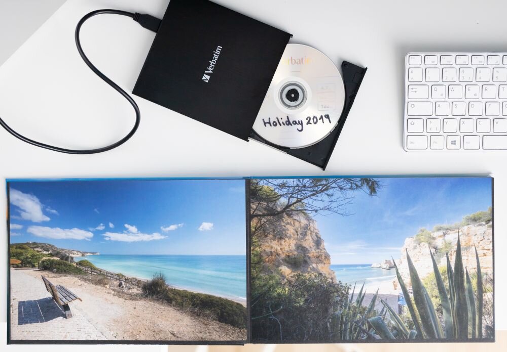 Nagrywarka Blu-ray VERBATIM (43889) Czarny napęd z wysuniętą tacką, w której znajduje się płyta z napisem 'Holiday 2019', obok klawiatura i otwarty album ze zdjęciami plaży i klifów lekka i zasilana przez USB-C