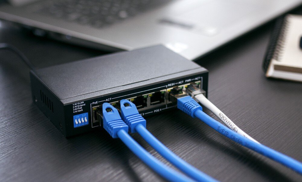 Switch EXTRALINK Krios EX-SG1005P Switch Krios na ciemnym biurku z trzema niebieskimi i jednym białym kablem sieciowym podłączonymi do portów